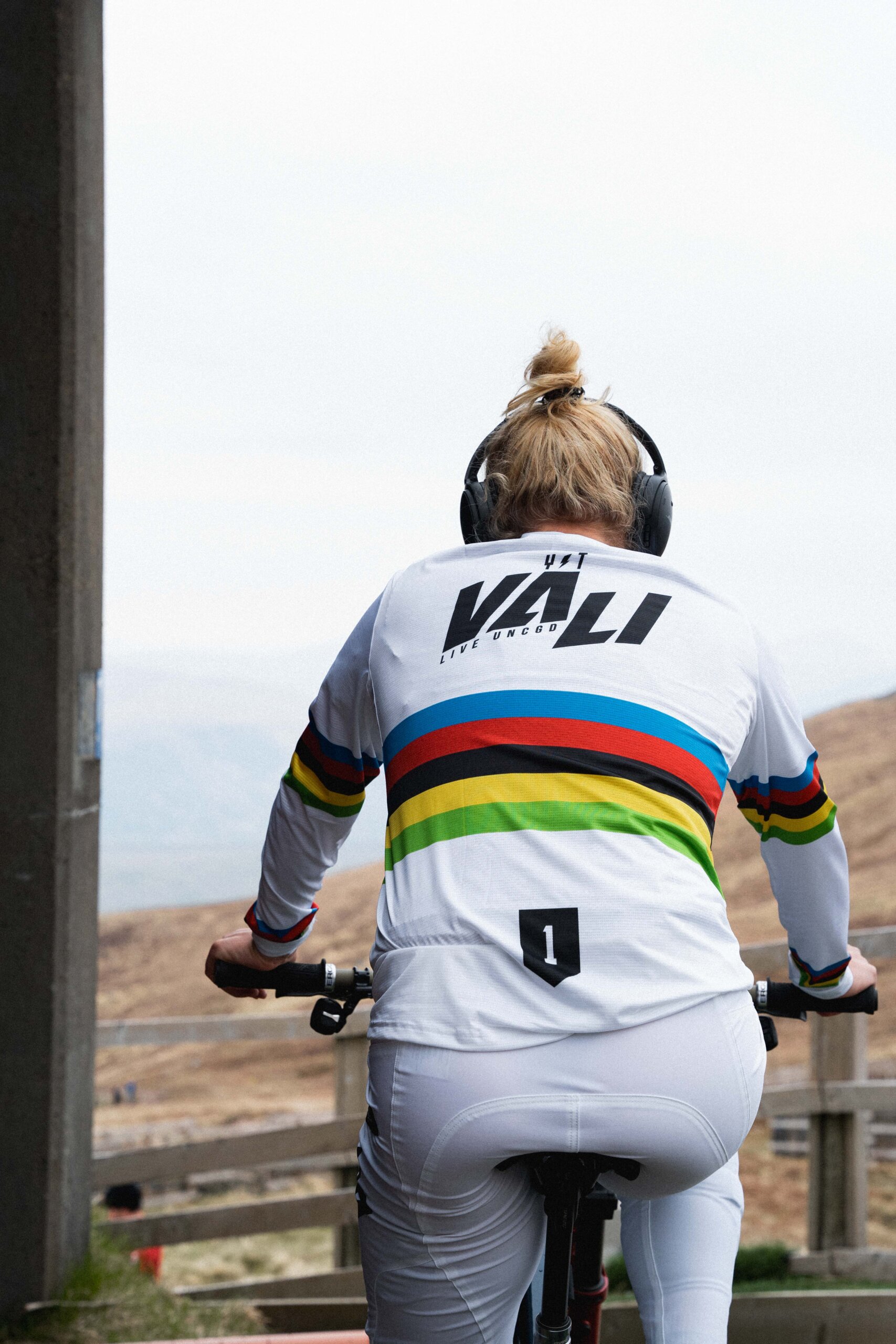Person auf Rad, Rücken, weißes Trikot mit bunten Streifen, Kopfhörer.